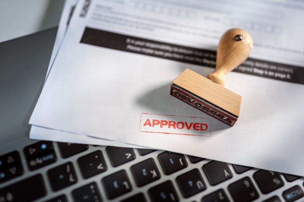 A red stamp that reads "Approved" on paper next to a computer keyboard.