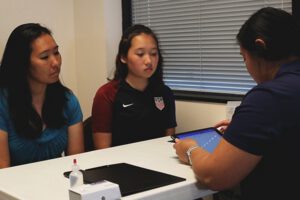 Young asian female athlete with mother talking to a doping control officer at a table.