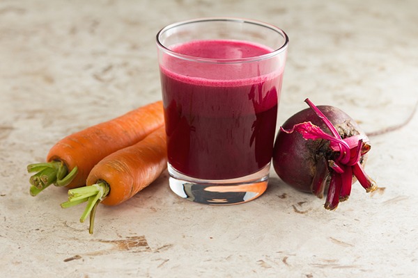 Glass of dark red liquid next to carrots and a beet.