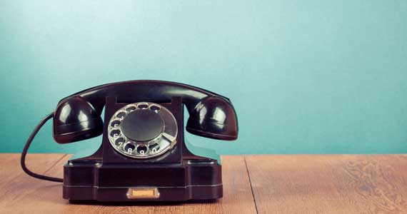 A black rotary phone against a blue background on a wood table.