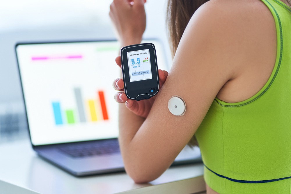 Woman with glucose monitor holding up tracking device and looking at laptop analytics.