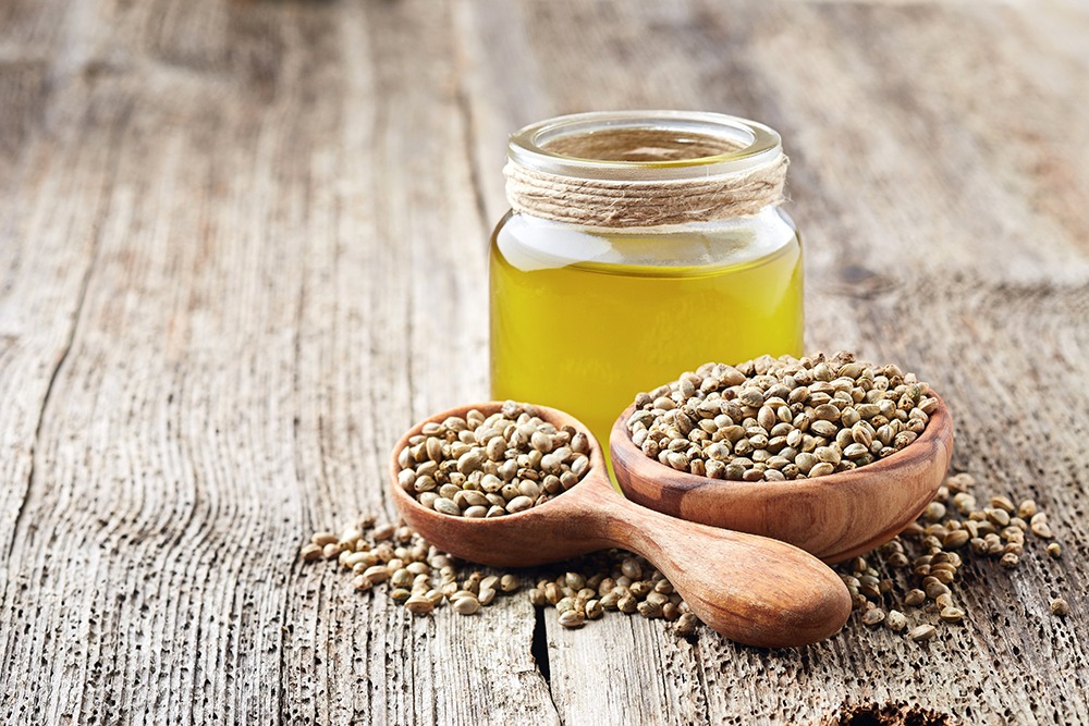 A bowl and spoon of hemp seeds next to a jar of hemp seed oil.