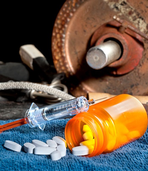 Pills falling out of an orange bottle with a syringe on top, next to a dumbbell.