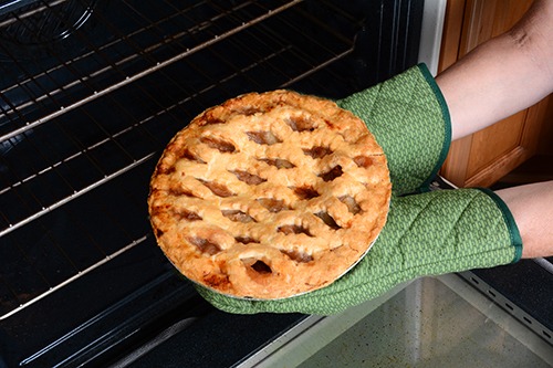 Close up of a pie coming out of an oven being held by a person wearing two green oven mitts.