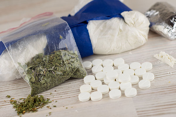 A table top with a bag of heroin, a clear bag of cocaine, a small bag of marijuana, and small, circular white pills.